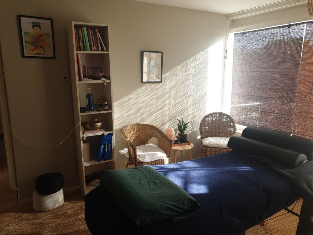 Een rustige behandelruimte met een massagetafel, een kast vol boeken en adviesmaterialen, en twee rieten stoelen bij een raam met zonlicht en schaduw.